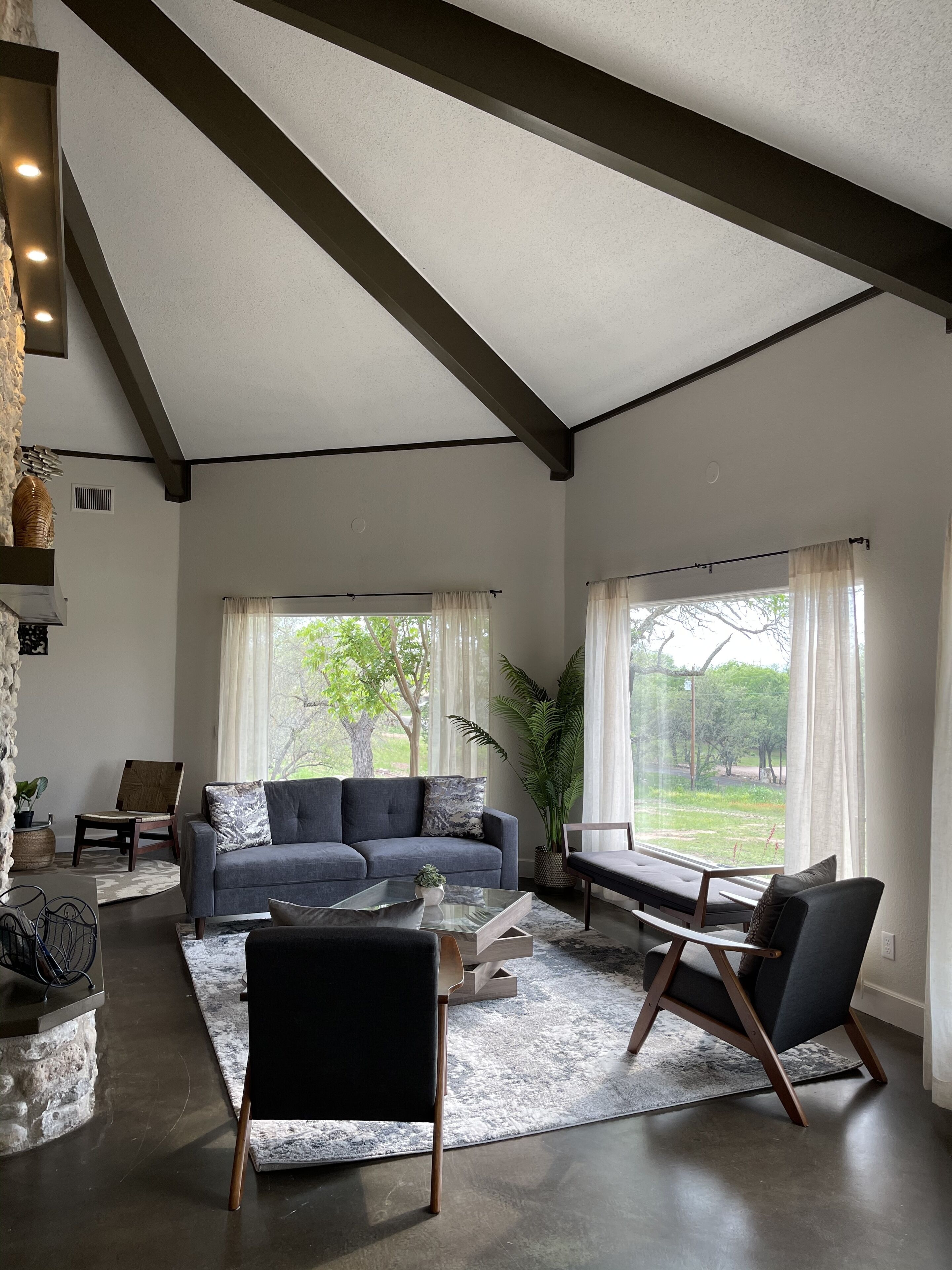 Ein modernes Wohnzimmer mit einem grauen Sofa, schwarzen Sesseln und einem Glastisch. Große Fenster mit leichten Vorhängen, eine Wand mit Steinverkleidung und ein Blick ins Grüne sind zu sehen.