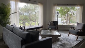 Uma sala de estar moderna com um sofá cinza, duas poltronas, uma mesa de centro de vidro e uma janela grande com cortinas leves.