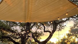 Uma área de refeições ao ar livre com uma mesa e cadeiras de madeira, um grande guarda-sol e vista para uma piscina.