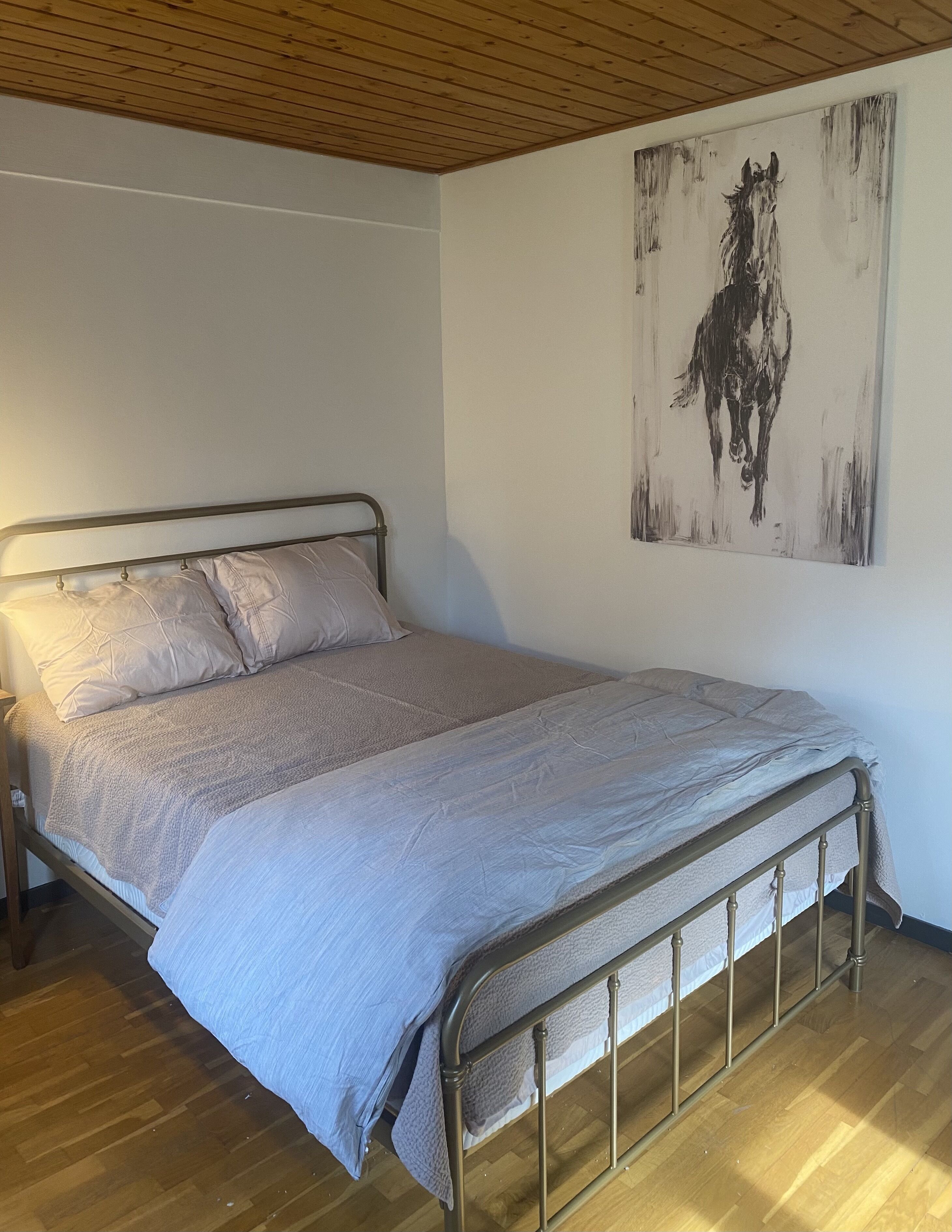 Ein Zimmer mit einem Einzelbett, einem Metallgestell, einer Holzbalkendecke und einem großen, abstrakten Gemälde an der Wand.