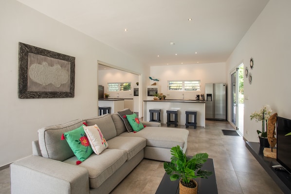 Ein modernes Wohnzimmer mit einem Ecksofa, einem Couchtisch und einem Fernseher. Im Hintergrund befindet sich eine Küche mit Kühlschrank, Backofen und Barhocker.