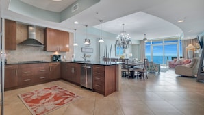 A spacious kitchen with modern cabinetry and a central island, a dining area with a chandelier, and a living room with a large window offering a view.