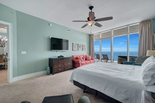 A bedroom with a large bed, a ceiling fan, a TV, a dresser, a red sofa, and a balcony with a view.