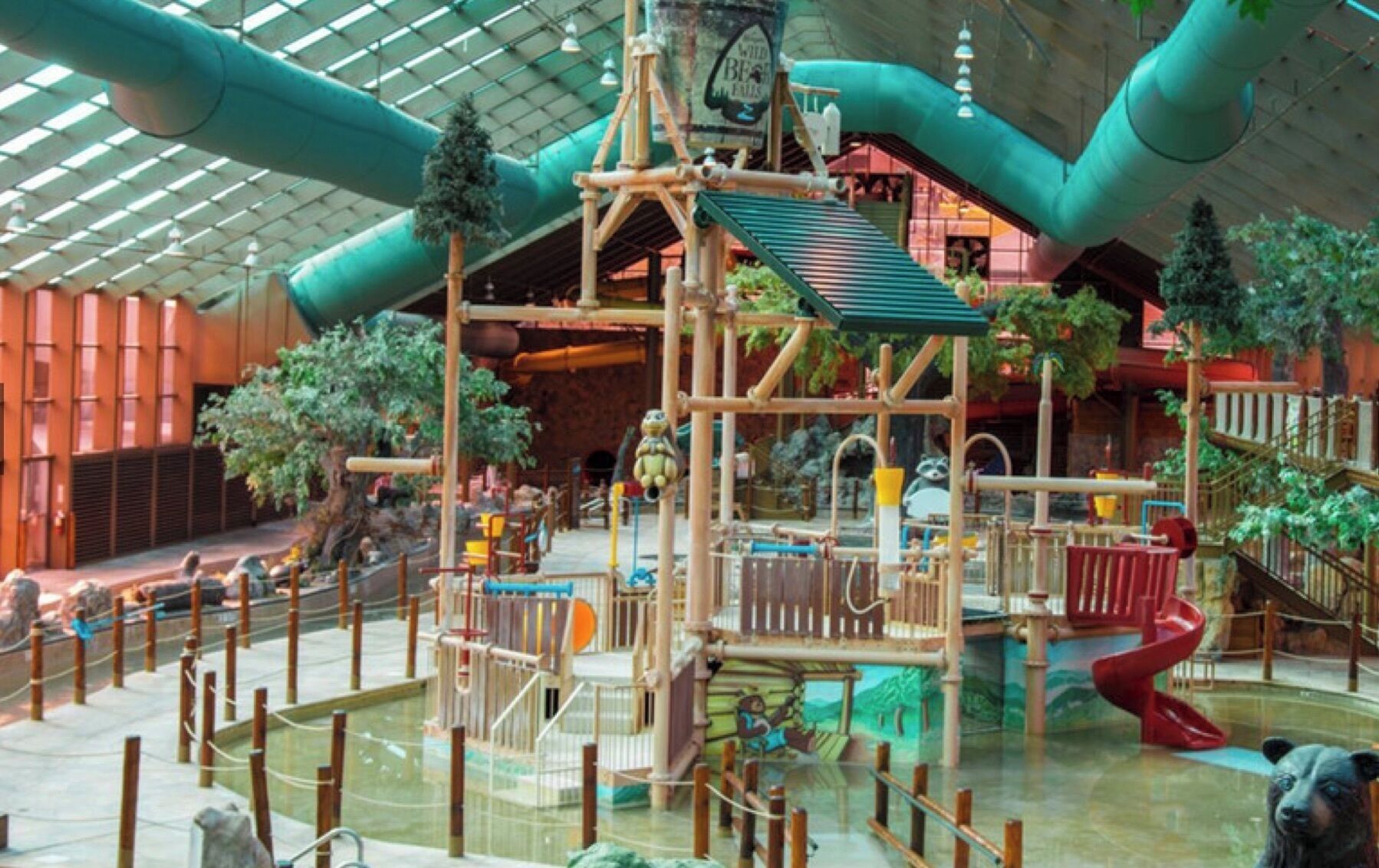 An indoor play area with a jungle theme, including a wooden climbing structure, slides, and artificial trees.