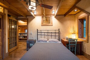 A bedroom with a wooden ceiling, a bed with a quilted bedspread, a desk with a chair, and a window with a view of trees. - Magical, stunning, secluded retreat on idyllic Lake Medora! (Copper Harbor)