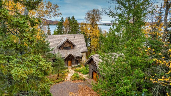 A rustic cabin with a stone exterior, a wooden door, and a chimney, surrounded by dense forest and a lake in the background. - Magical, stunning, secluded retreat on idyllic Lake Medora! (Copper Harbor)