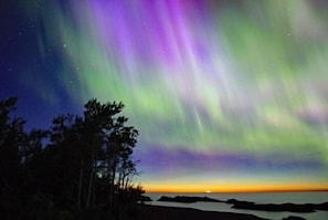 A night sky with the Northern Lights over a coastal landscape with trees and water. - Magical, stunning, secluded retreat on idyllic Lake Medora! (Copper Harbor)