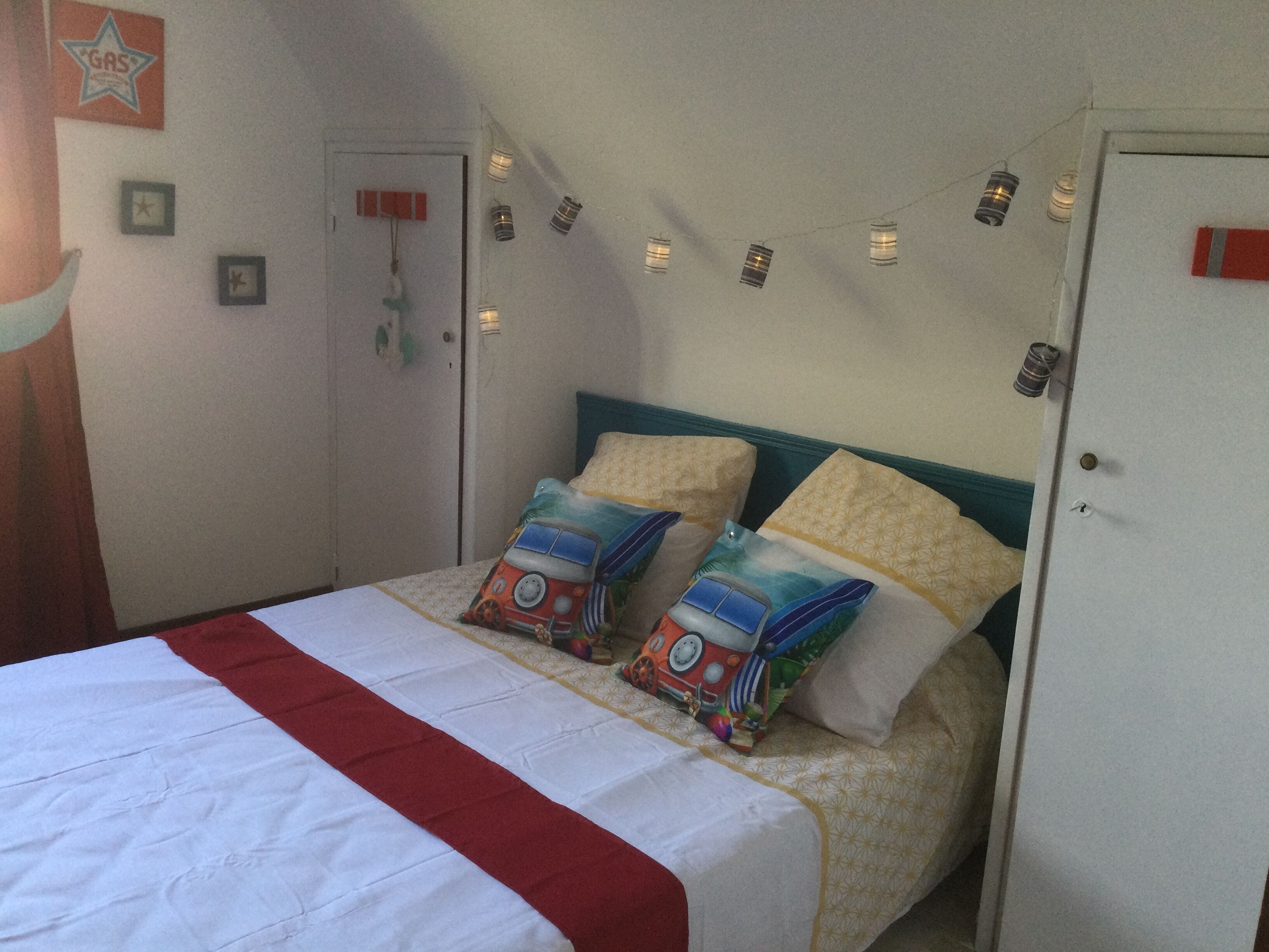 A neatly made bed with a red and white bedspread, two pillows with a colorful design, and string lights on the wall.