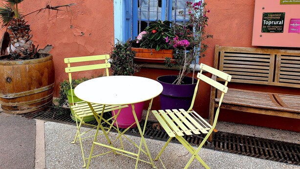 A small outdoor seating area with a round table and two yellow chairs, a wooden barrel, and a potted plant with pink flowers. - Gîte T2 Aspres (Tautavel)