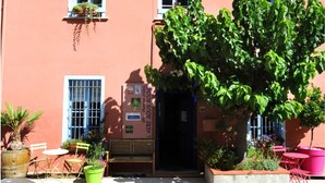 A building with a pink facade, a large tree in the center, and outdoor seating with chairs and tables. - Gîte T2 Aspres (Tautavel)