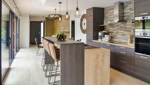 A modern kitchen with a central island, bar stools, a built-in oven, and a large clock on the wall. - Villa Cosy, Pool, Spa, Relaxation Area (Sauna & Hammam) 8 km from the beaches! (Pordic)