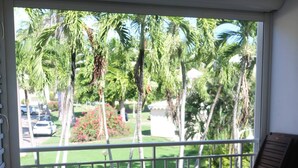 A balcony with a table and chairs, overlooking a garden with palm trees and a parked car.