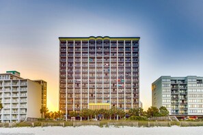 A high-rise hotel with multiple balconies, located near a beach with a sandy foreground.