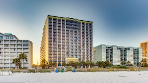 Hotel alto com várias varandas, situado numa praia com palmeiras e areia à frente.