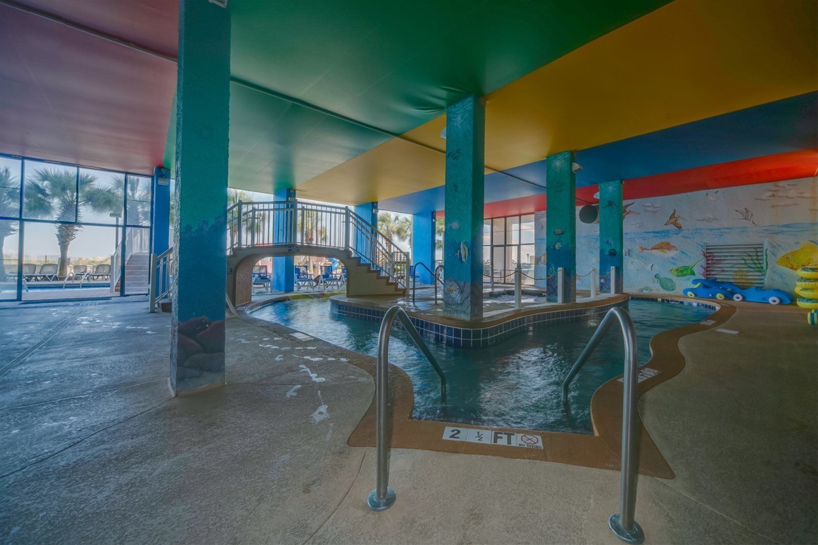 Piscina interna com uma ponte, rodeada por paredes coloridas e vista para palmeiras.