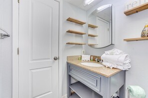 A bathroom with a white door, a sink with a granite countertop, a mirror, and wooden shelves above the mirror.