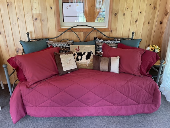 A bed with a red quilted cover, wooden headboard, and multiple decorative pillows. - Knotty Pine - Private cabin nestled in the woods, fire pit, near CDP/ASV (Hartwick)