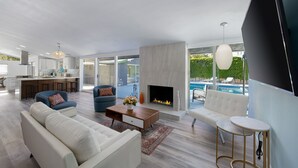 Una sala de estar moderna con chimenea, vista a la piscina y una cocina al fondo.