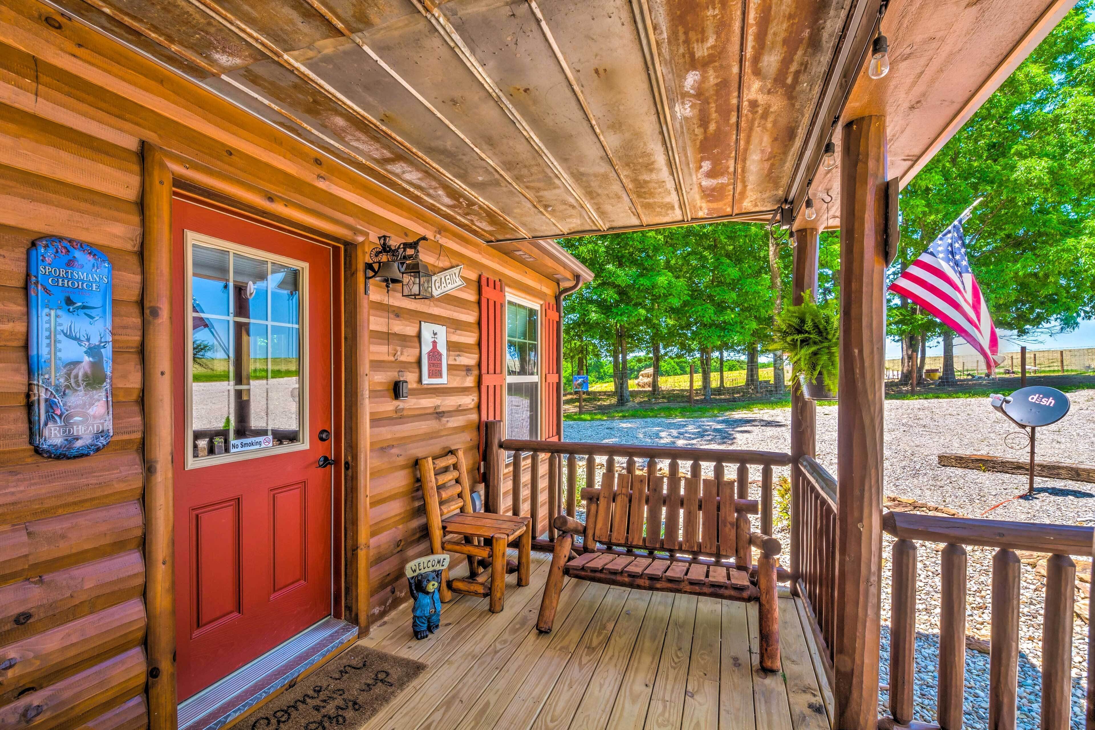 Een houten chalet met een rode deur, een veranda met een bankje, een welkomstbord en een Amerikaanse vlag.