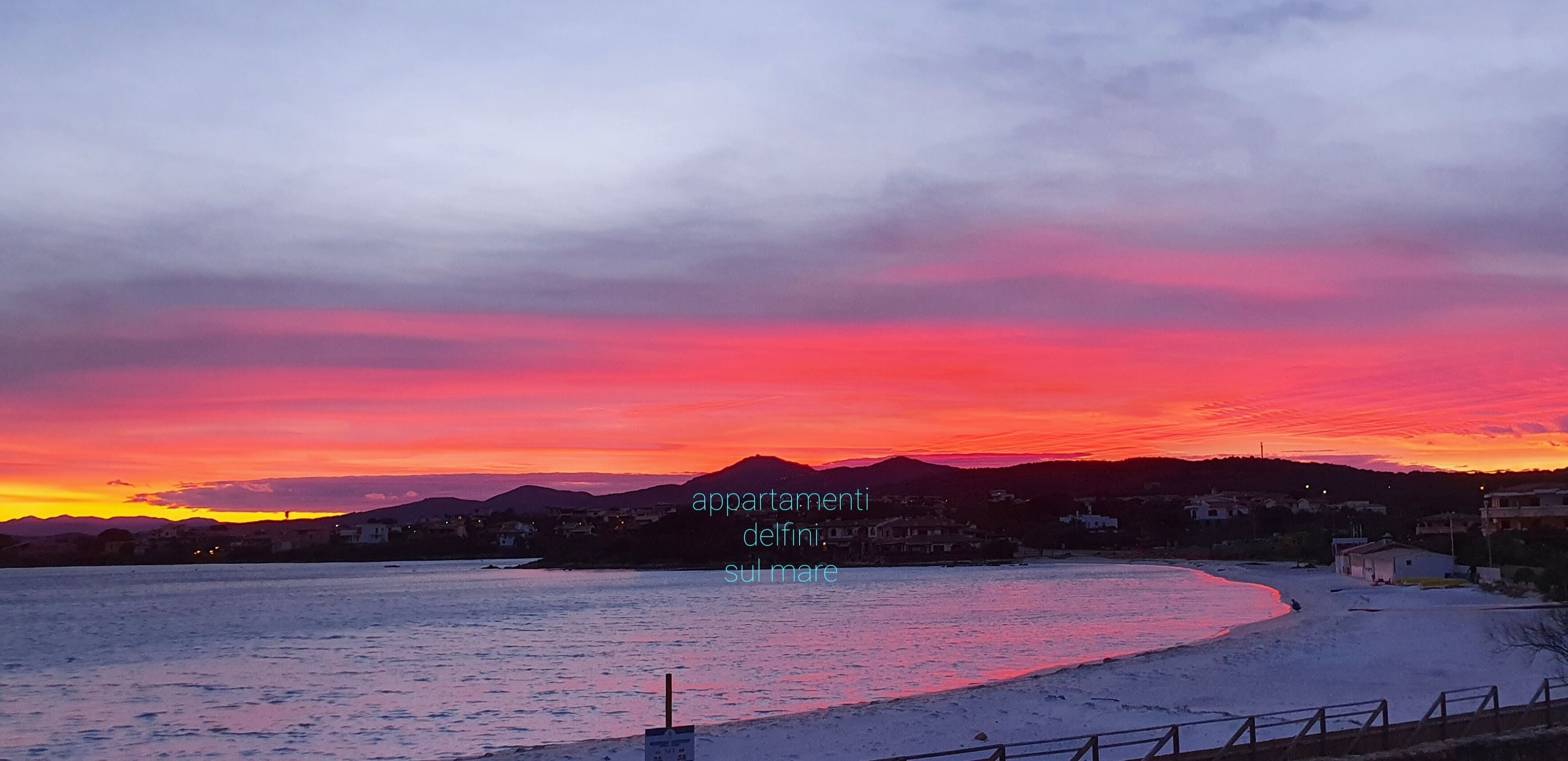 A coastal landscape with a sunset, a beach, and a row of buildings in the background.