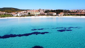 Blick auf die Küste mit klarem türkisblauem Wasser, einem Sandstrand und einem dicht bebauten Wohngebiet mit mehrstöckigen Gebäuden.