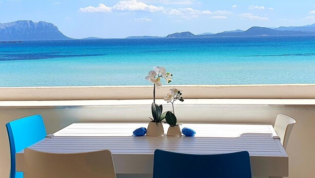 Ein Balkon mit Tisch und Stühlen mit Blick auf ein klares, blaues Meer und ferne Berge.