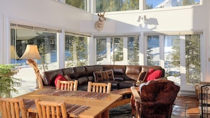 A modern interior with a leather sofa, wooden dining table, and chairs, large windows with a mountain view, and a mounted animal head. - Ski in/out home w/private hot tub, sauna, fireplace & great views - Low Dog Mountain Chalet (Big Sky)