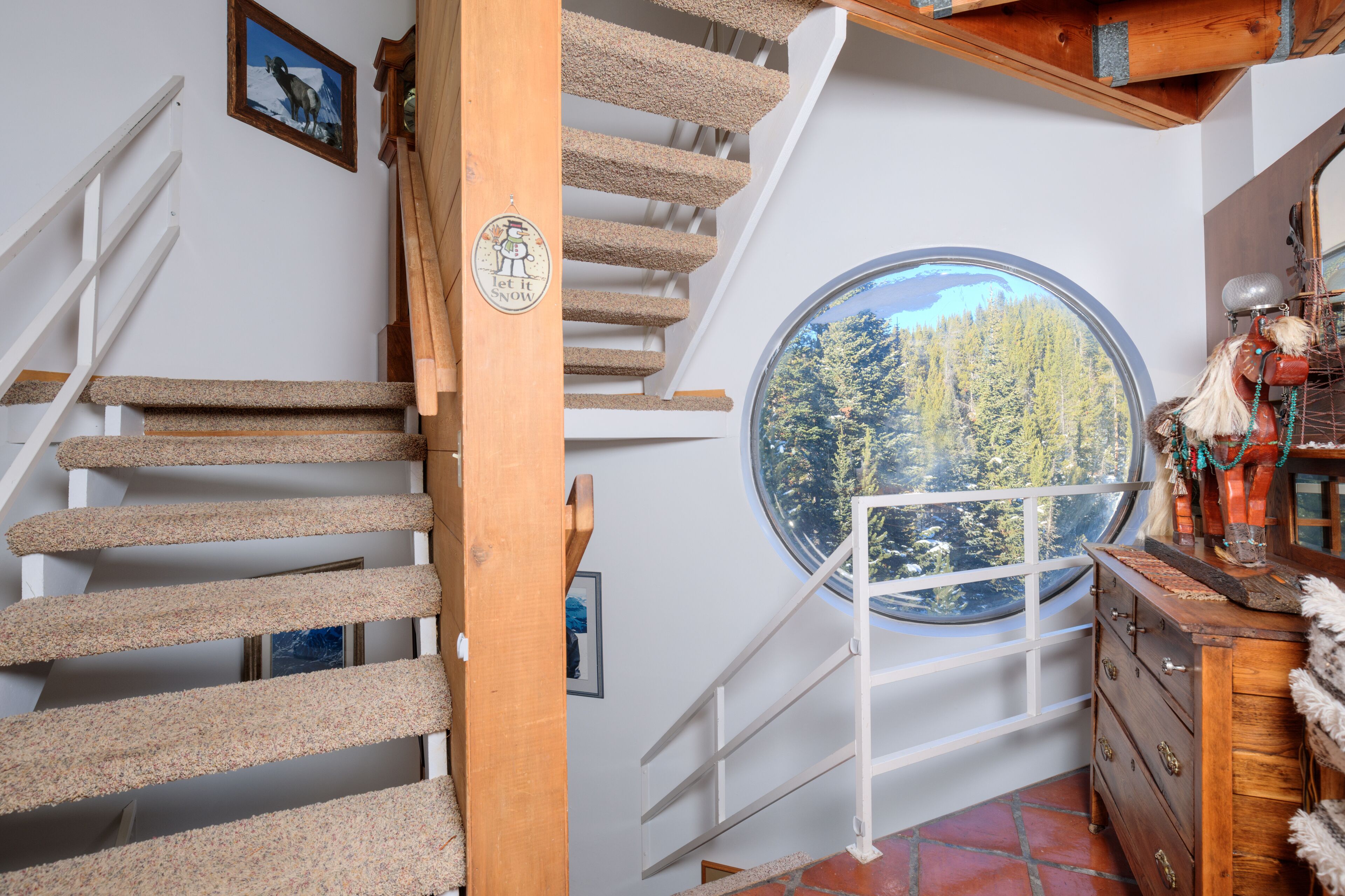 Eine Wendeltreppe mit Holzgeländer und Teppichstufen führt zu einem Raum mit einem großen Rundfenster, das einen Blick auf einen Wald bietet.