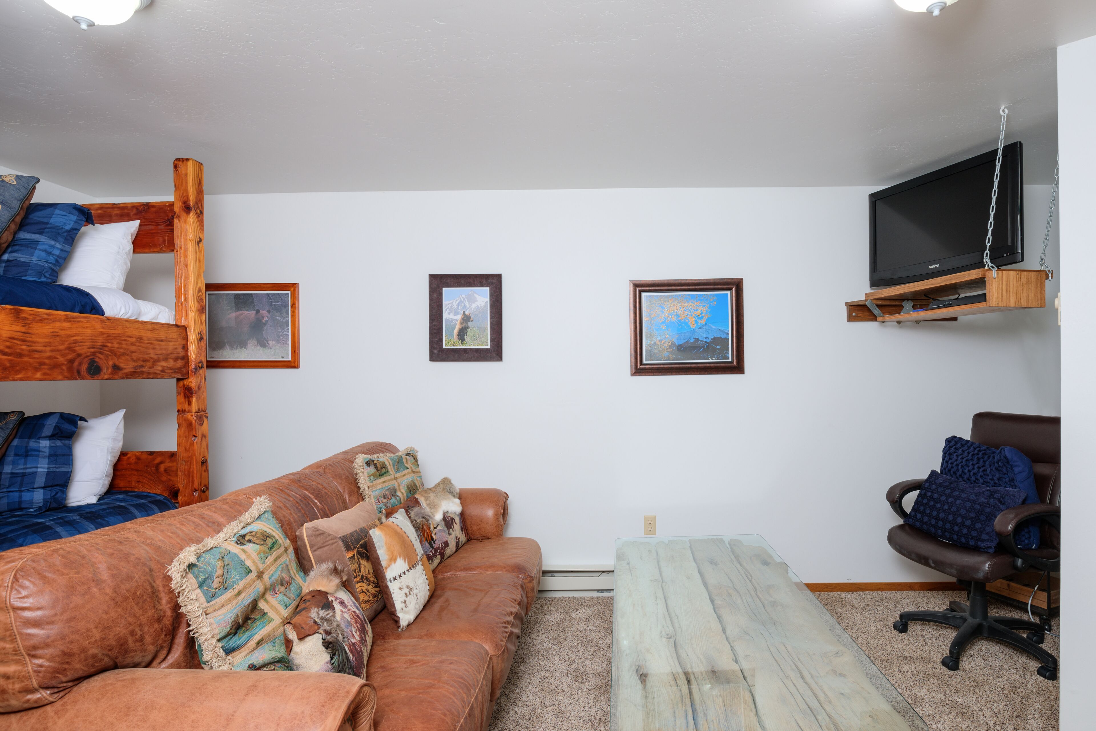 Una habitación con una cama nido, un sofá, una mesita de centro, una silla y un televisor montado en la pared.