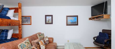 Una habitación con una cama nido, un sofá, una mesita de centro, una silla y un televisor montado en la pared.