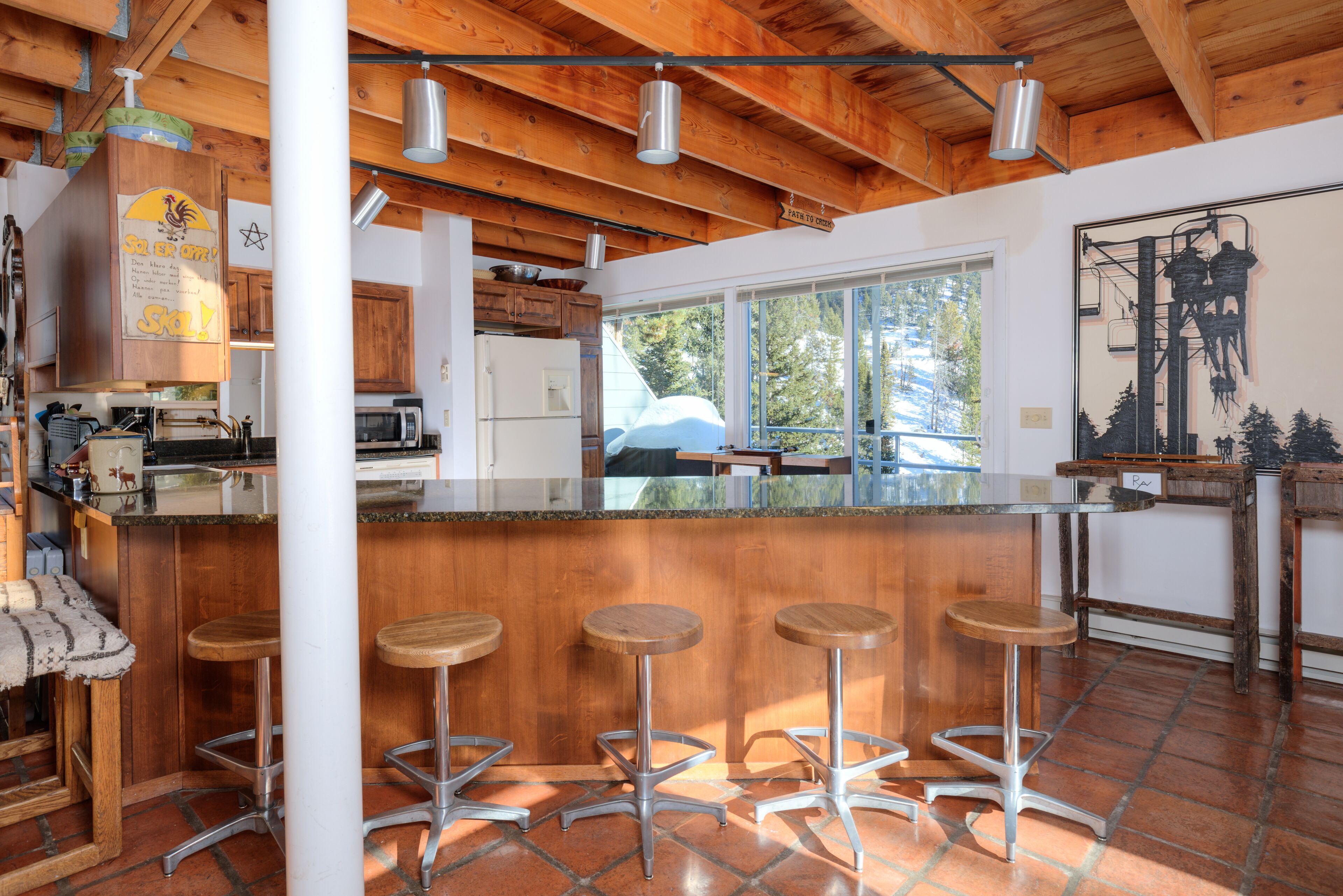 A kitchen with wooden beams, a large island with stools, and a view of the outdoors.