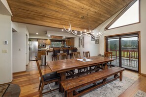 A spacious dining area with a wooden table and benches, a kitchen in the background, and a view through a large sliding glass door.