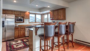 A kitchen with wooden cabinets, a stainless steel refrigerator, a microwave, and a large island with bar stools. - Private Hot Tub | Golf Course | Nordic Skiing | Big Sky Awaits (Big Sky)