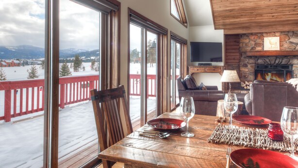 A dining area with a table set for four, large windows, a stone fireplace, and a view of a snowy landscape. - Private Hot Tub | Golf Course | Nordic Skiing | Big Sky Awaits (Big Sky)