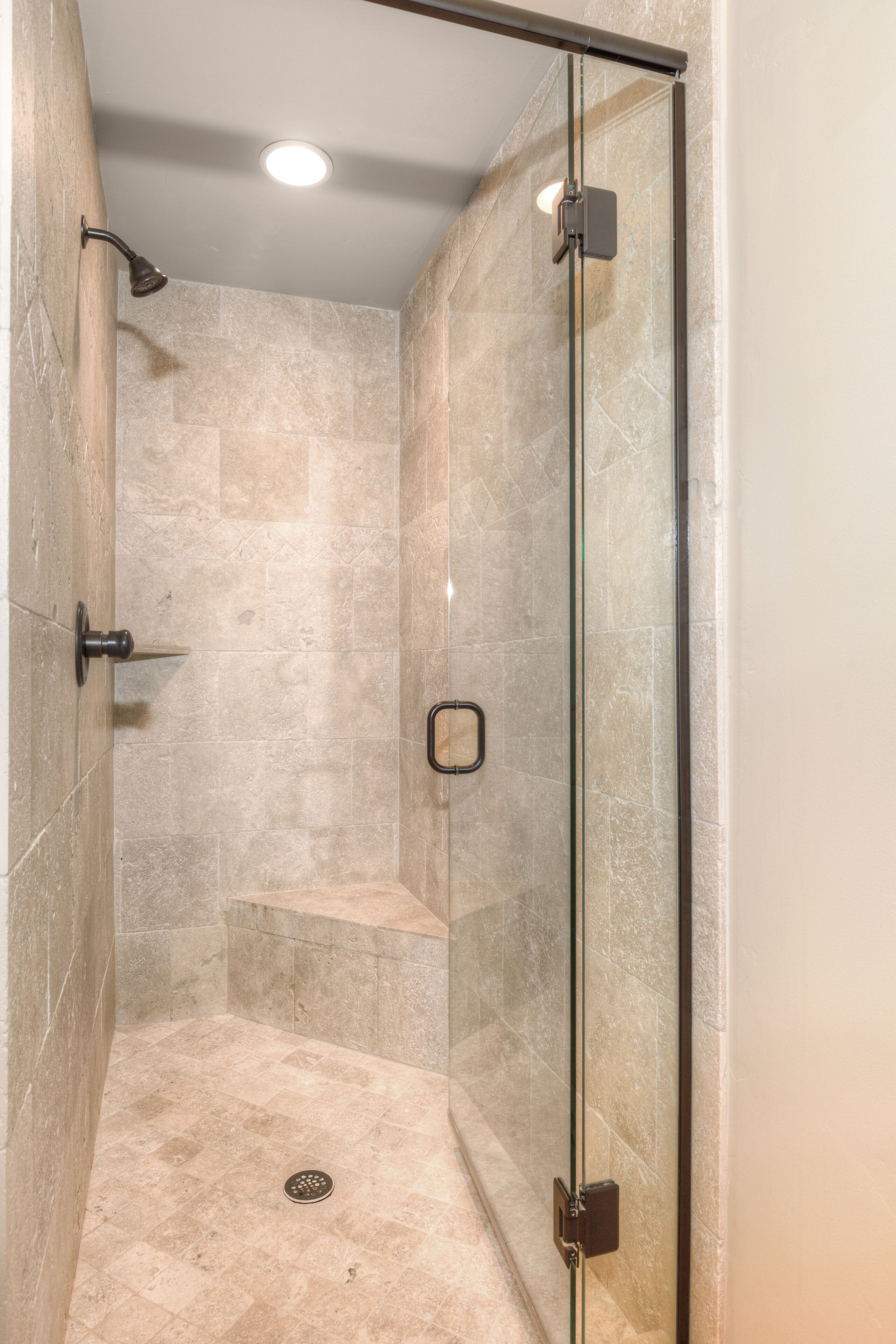 A glass-enclosed shower stall with beige tiles, a showerhead, and a small shelf.