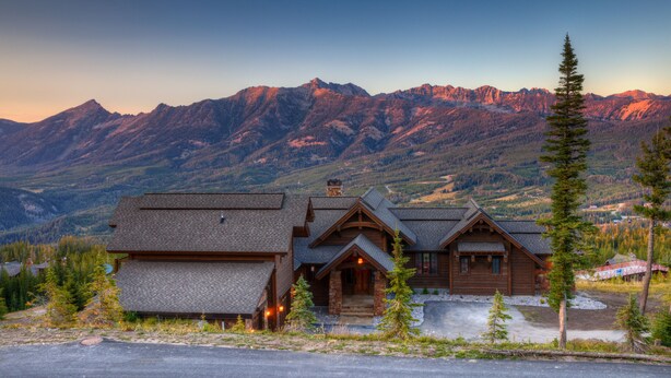 Une cabane en rondins, avec des fondations en pierre, un revêtement extérieur en bois et un porche à l’avant. Elle est entourée d’une zone boisée, avec des montagnes en arrière-plan.