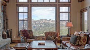 Un salon spacieux doté de grandes fenêtres offrant une vue sur les montagnes, de meubles en cuir et d’une cheminée.