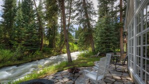 A stone patio with a fire pit, white chairs, and a view of a river and forest. - Secluded home close to Meadow Village w/private hot tub & library | Creekside Retreat (Big Sky)