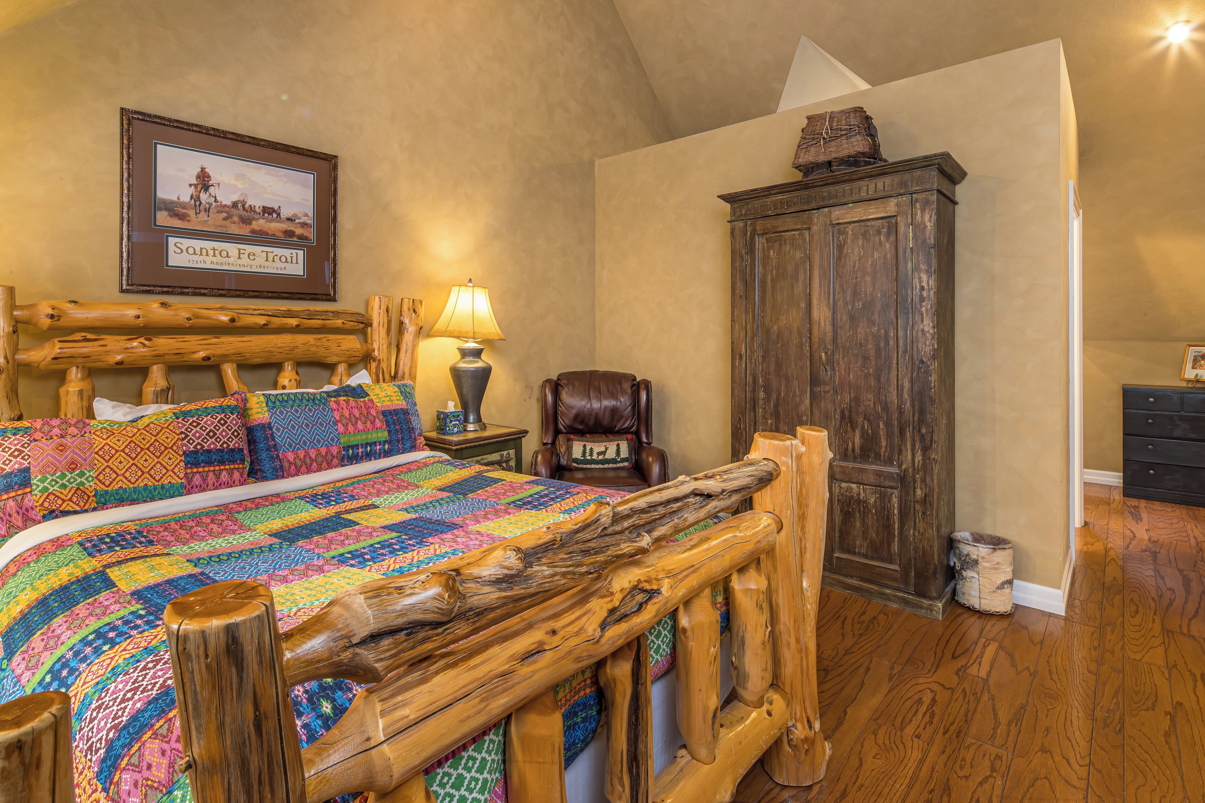 Une chambre à coucher avec un cadre de lit en bois, une courtepointe aux couleurs vives, un tableau encadré représentant le sentier de Santa Fe, une armoire en bois et une lampe.