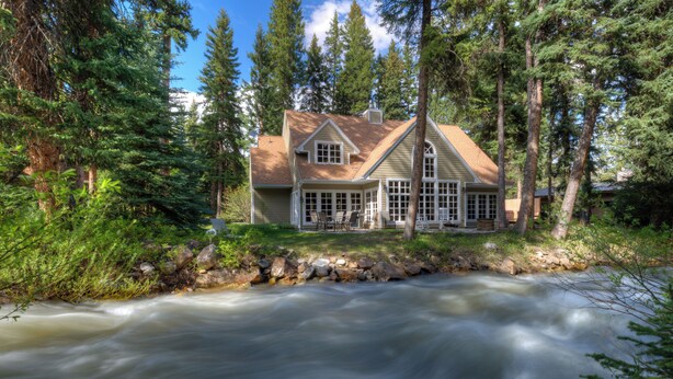A house with a porch overlooking a river, surrounded by trees. - Secluded home close to Meadow Village w/private hot tub & library | Creekside Retreat (Big Sky)