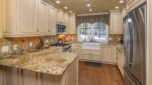 A kitchen with wooden cabinets, a granite countertop, a stainless steel refrigerator, and a window with a view of trees. - Secluded home close to Meadow Village w/private hot tub & library | Creekside Retreat (Big Sky)