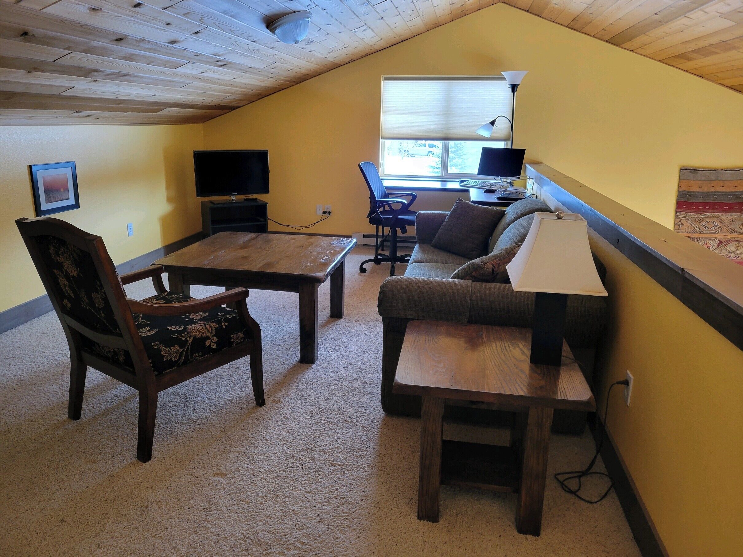 A cozy, well-lit room with a wooden ceiling, a sofa, a desk, a chair, and a television.