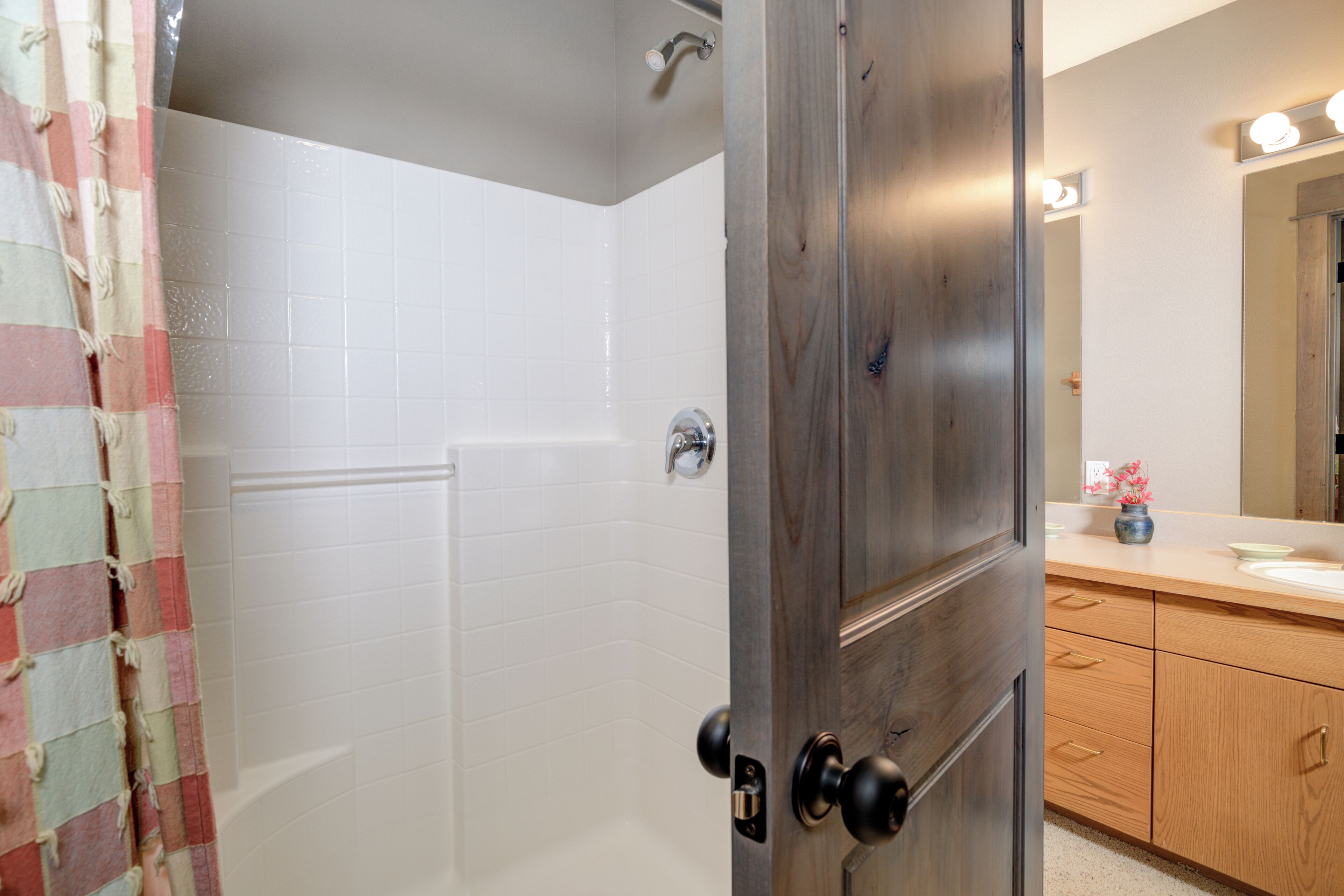 Une salle de bain avec des murs recouverts de carreaux blancs, une porte en bois, une douche et un meuble-lavabo avec un miroir.