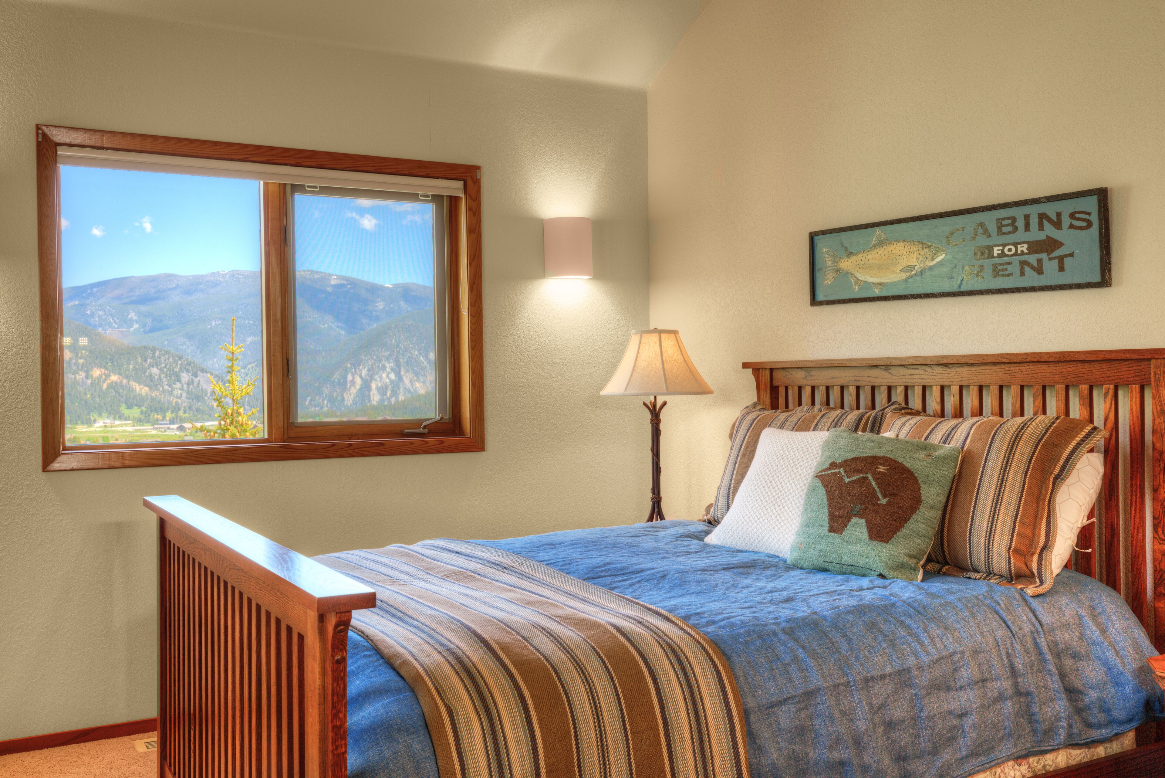 A bedroom with a large window, a bed with a striped blanket, a lamp, and a wall sign indicating cabins for rent.