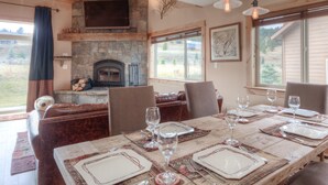 Un espace repas agrémenté d’une cheminée en pierre, d’un plafond en bois et de grandes fenêtres donnant sur l’extérieur.