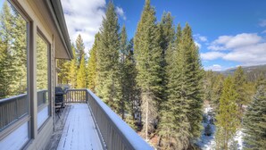 A wooden deck with a railing overlooking a forested area with snow patches. - Home in Big Sky Town Center w/fireplace & community pool/hot tub - Hidden Gem in the Trees (Big Sky)
