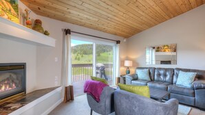 Una sala de estar con chimenea, un conjunto de sofá gris, techo de madera y vista a un paisaje verde a través de un ventanal corredero.