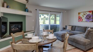 Un coin repas avec une table ronde dressée pour quatre personnes, entourée de chaises en bois. On aperçoit un canapé et un téléviseur à l’arrière-plan.