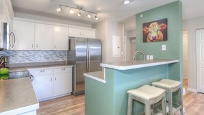 Une cuisine moderne avec des meubles blancs, un réfrigérateur en acier inoxydable, un mur d’accent de couleur turquoise et deux tabourets de bar.