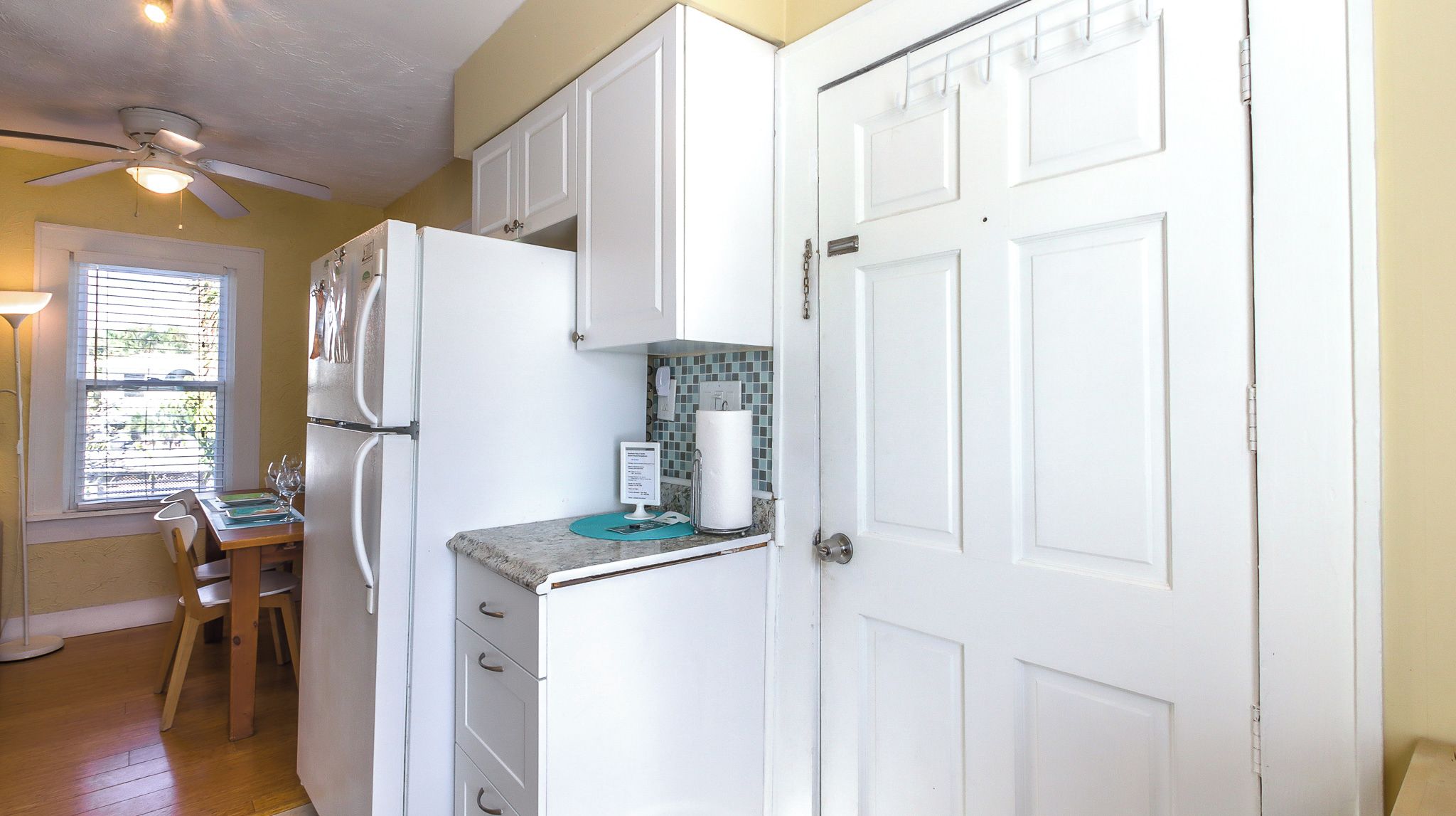 Una cocina compacta con armarios blancos, un refrigerador, una encimera con dispensador de toallas de papel y una ventana con persianas.